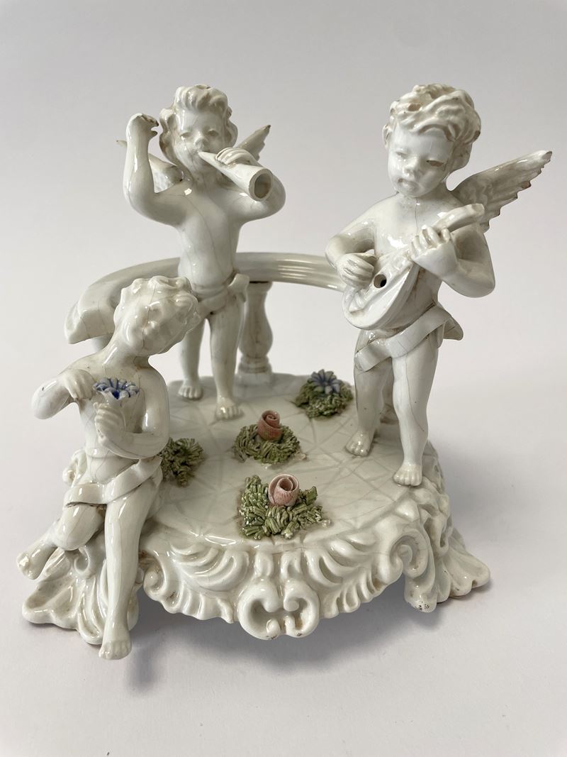 Composizione in porcellana bianca con inserti policromi di fiori raffigurante tre putti che suona...