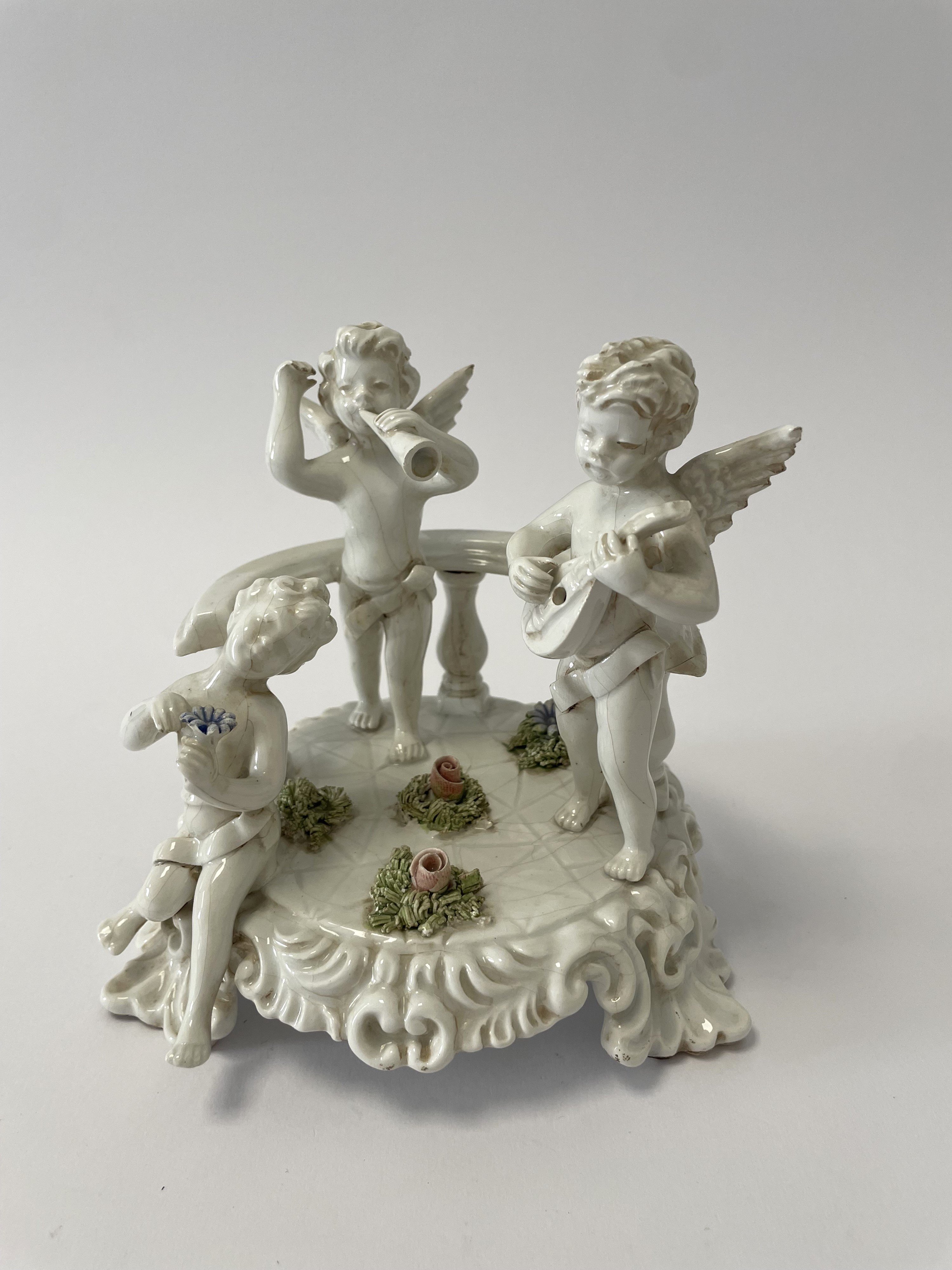Composizione in porcellana bianca con inserti policromi di fiori raffigurante tre putti che suona...