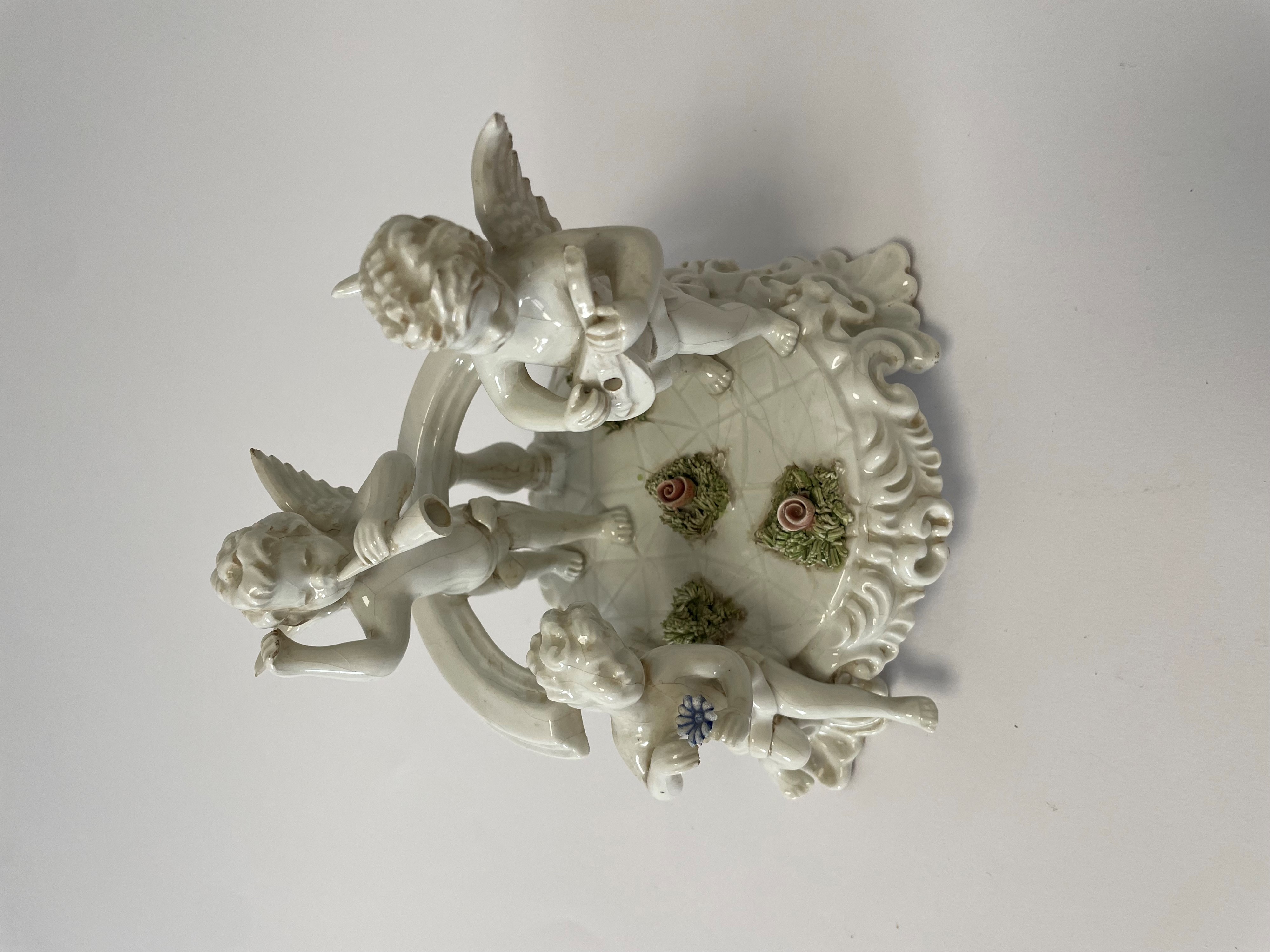 Composizione in porcellana bianca con inserti policromi di fiori raffigurante tre putti che suona...