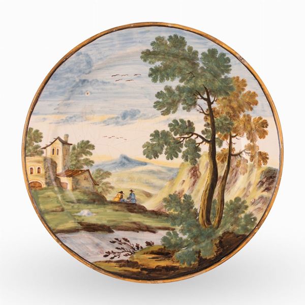 Manifattura castellana del XVIII secolo - Piatto in maiolica decorato in policromia con paesaggio alberato, fiume, personaggi e case