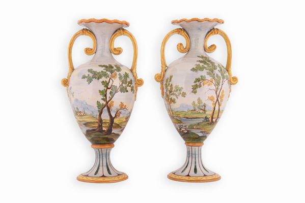 Manifattura castellana del XX secolo - Coppia di vasi biansati in maiolica con corpo ad anfora su alto piede, decorai in policromia con paesaggi alberati e case. Bordo superiore ondulato.