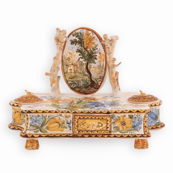 Manifattura Antonio de Simone, Castelli XX secolo - Calamaio in maiolica a fondo bianco decorato in policromia. Al centro medaglione ovale con paesaggio sorretto da putti.