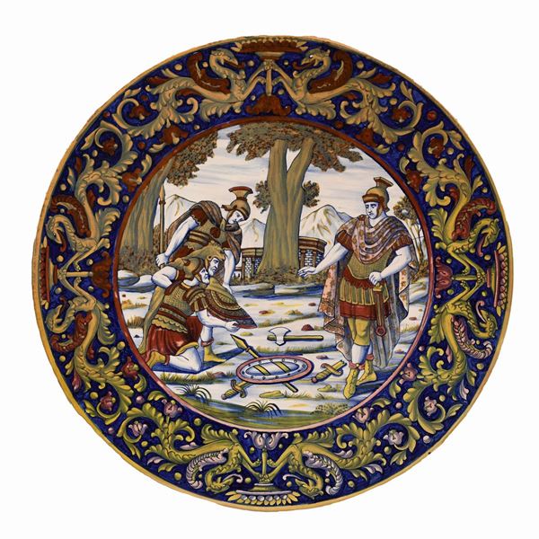 Grande piatto Gualdo Tadino dipinto a lustro. Tesa dipinta a grottesche e girali fogliati. Cavetto reca la raffigurazione di una resa militare con soldati romani. Siglato al retro SCU Italy P.R. (Paolo Rubboli)