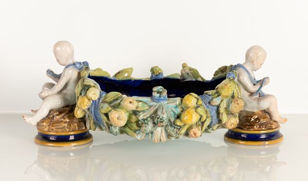 Centrotavola di gusto robbiano neoquattrocentesco con ai lati due putti in maiolica bianca modellati a tutt tondo che trattengono una ghirlanda di frutta e fiori formante le pareti dell'oggetto dipinto all'interno in blu di Prussia