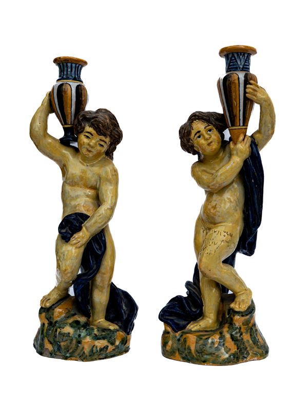 Coppia di putti tedofori in maiolica stanti a trattenere due anfore 