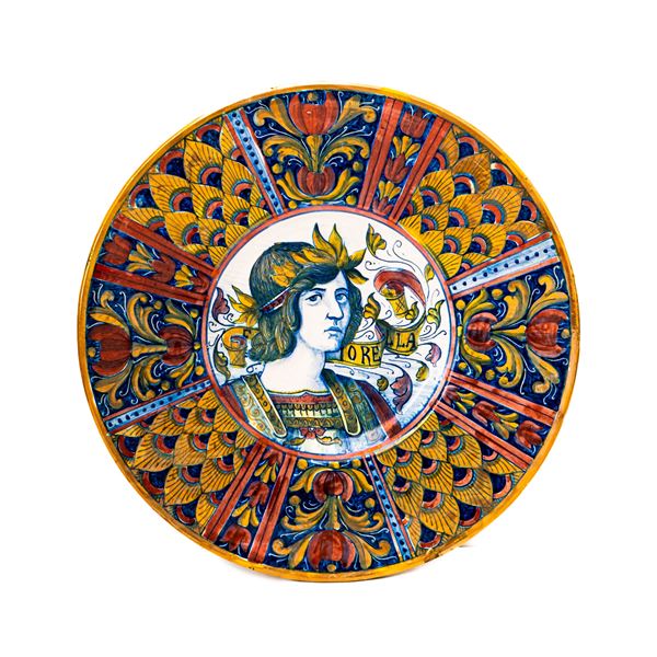 Manifattura di Gualdo Tadino del XX secolo - Piatto in maiolica decorato a lustro metallico. Nella tesa si alternano decori a penna di pavone con altri a fiori fantastici. Nel cavetto figura muliebre e cartiglio " Fiorella". S.M.G ( Società Mastro Giorgio)