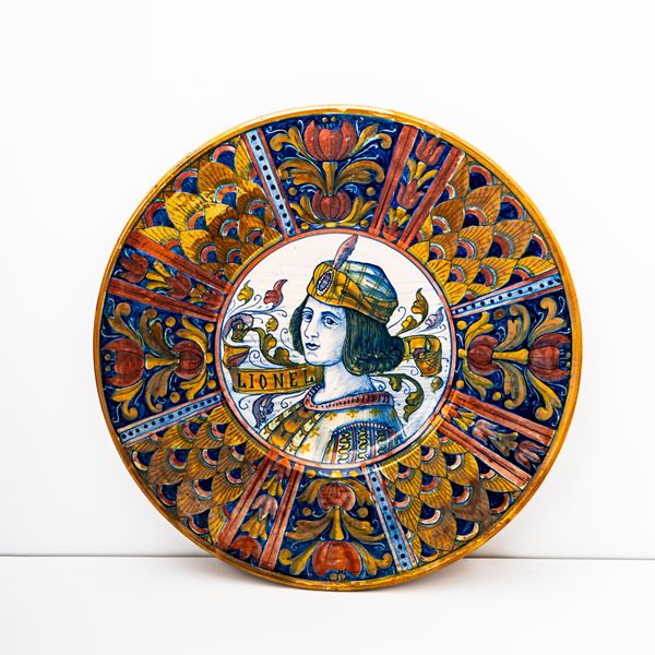 Manifattura di Gualdo Tadino del XX secolo - Piatto in maiolica decorato a lustro metallico. Nella tesa si alternano decori a penna di pavone con alri a fiori fantastici. Nel cavetto figura muliebre e cartiglio "Lionello". S.M.G ( Società Mastro Giorgio)