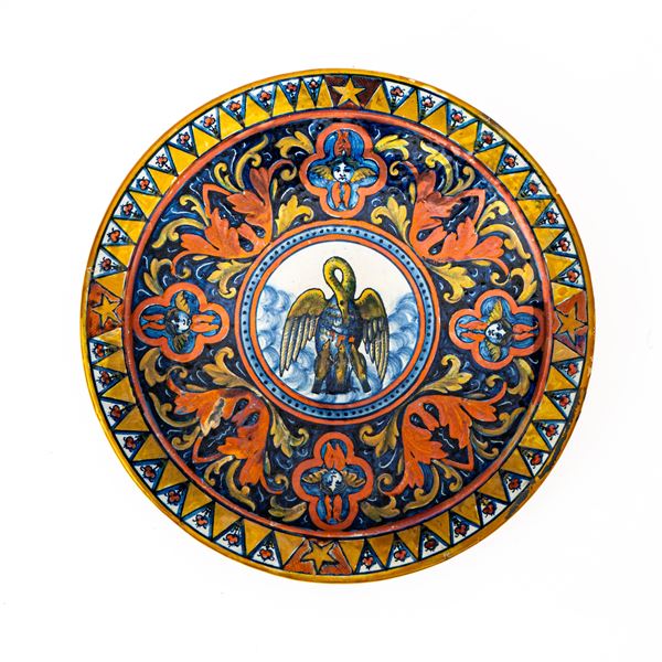 Manifattura di Gualdo Tadino del XX secolo - Piatto in maiolica decorato a lustro metallico. Nella tesa decoro alternato di triangoli con quattro stelle ai punti cardinali. Quattro mascheroni alternati a decori floreali incorniciano un tondo con decoro del pellicano che sfama i piccoli beccandosi il petto. 